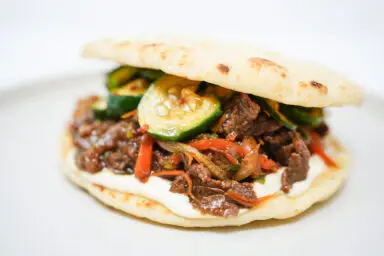 A Beef Bulgogi Sandwich on toasted naan bread, topped with spicy marinated cucumbers, a drizzle of garlic mayo, and fresh cilantro leaves, showcasing a delicious fusion of flavors.
