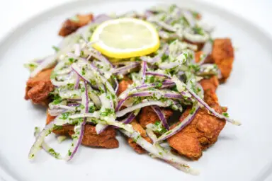 Golden, crispy fried pork belly slices on a plate, accompanied by a zesty onion and parsley salad, offering a refreshing contrast to the rich, savory pork, ready for a flavorful dining experience.