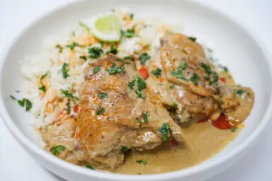 A vibrant dish of Mango Coconut Braised Chicken Thighs, featuring juicy, golden-brown chicken thighs in a creamy, tropical mango and coconut sauce, garnished with fresh cilantro on a white plate.