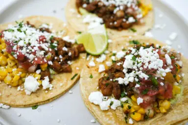 A plate of Ancho Chili Chicken Tacos featuring tender, spiced chicken pieces, golden charred corn, and vibrant tomato salsa, all tucked into soft corn tortillas and garnished with crumbled cheese and fresh cilantro.