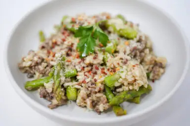 A creamy Arborio rice risotto enriched with savory Italian sausage and tender green asparagus, garnished with red pepper flakes and fresh parsley, served in a white bowl for a comforting and colorful meal.