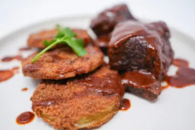 A plate of Southern-Inspired Braised Beef Short Ribs and Fried Green Tomatoes, showcasing tender, slow-cooked ribs draped in a rich, glossy sauce alongside golden-brown, crispy fried green tomatoes, embodying a hearty and flavorful Southern meal.