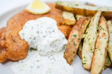 A plate of golden, crispy beer-battered fish alongside oven-baked garlic parsley potato wedges, served with a side of creamy homemade tartar sauce.