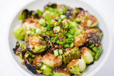 A colorful plate of roasted Brussels sprouts drizzled with peanut sauce, topped with green onions and red pepper flakes.