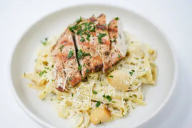 A plate of Grilled Chicken with Roasted Garlic Fettuccine Alfredo, featuring golden grilled chicken slices atop creamy, white Alfredo pasta sprinkled with fresh green parsley.
