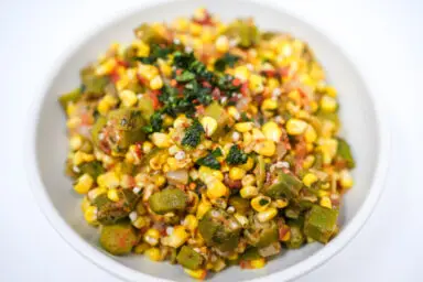 A vibrant dish of Corn and Okra Succotash, featuring bright yellow corn kernels and green okra slices mixed with red bell pepper and diced tomatoes, seasoned with paprika and chili, and garnished with fresh basil, served in a rustic bowl.