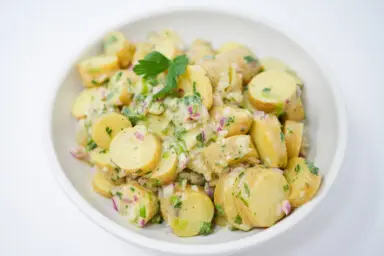 A colorful bowl of Tangy Fingerling Potato, Mustard, and Onion Salad, featuring sliced fingerling potatoes, red and green onions, drizzled with a golden mustard vinaigrette and sprinkled with fresh chopped parsley.