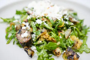 A vibrant image of Grilled Sausage and Summer Squash Salad featuring slices of smoky grilled Italian sausage, charred zucchini and yellow squash, and crisp arugula leaves, all tossed together and sprinkled with crumbled goat cheese. The salad is drizzled with a lemon herb vinaigrette and garnished with red pepper flakes, adding a subtle hint of spice. The colors and textures create a visually appealing and appetizing dish.