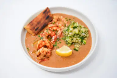 A colorful plate of Grilled Royal Red Shrimp over vibrant heirloom tomato gazpacho, accompanied by a fresh grilled zucchini salsa and a side of lightly charred naan bread, showcasing a harmonious blend of fresh summer flavors and textures.