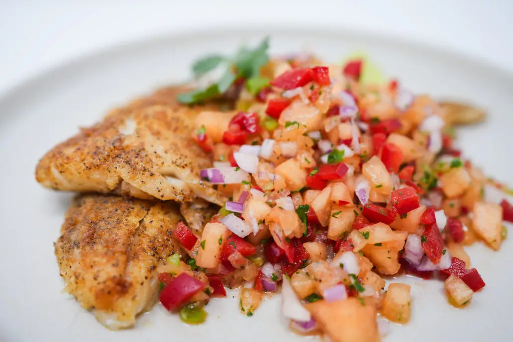 A vibrant close-up of cantaloupe salsa aside a pan-fried haddock fillet, showcasing the bright orange cantaloupe chunks mixed with red bell pepper and cilantro, adding a colorful contrast to the golden-brown fish.






