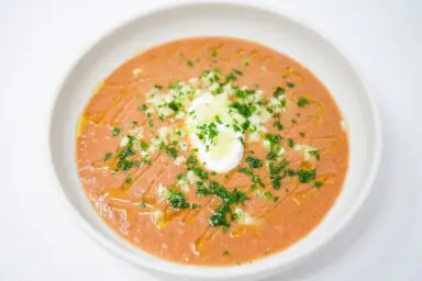 A bowl of Heirloom Tomato Gazpacho, featuring a vibrant red tomato base with chunks of cucumber and a dollop of white sour cream on top, garnished with fresh green parsley.
