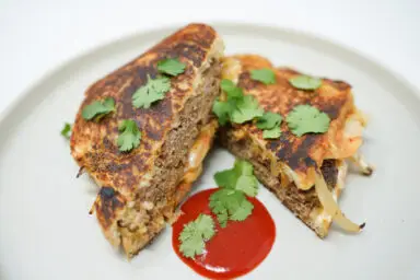 A Korean BBQ Patty Melt on a plate, showcasing a juicy beef patty with caramelized onions and kimchi, melted cheese between toasted sourdough slices, served with a side of sriracha sauce for dipping.