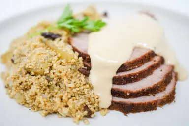 A succulent, spice-crusted eye round roast beef, sliced and generously drizzled with creamy lemon-tahini sauce, accompanied by a fluffy side of couscous, all presented on a white platter showcasing Moroccan-inspired culinary elegance.