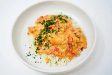 A serving of Passionfruit Coconut Chicken featuring golden-brown chicken thighs nestled in a creamy, pale orange sauce made from passionfruit and coconut milk, garnished with vibrant green cilantro, served over a bed of fluffy white basmati rice.