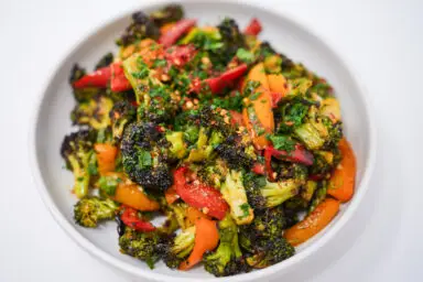A colorful dish of Spicy Peanut Roasted Broccoli and Bell Peppers, featuring vibrant red and orange bell peppers and deep green broccoli florets, all coated in a glossy, spicy peanut sauce and sprinkled with fiery red pepper flakes.
