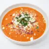 A bowl of Roasted Tomato, Cantaloupe, and Pancetta Soup, featuring a creamy blend of vibrant orange soup topped with golden crispy pancetta, crumbled goat cheese, and a sprinkle of fresh green parsley.