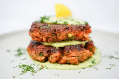 A plate of Salmon Cakes with Avocado Dill Cream features golden-brown salmon patties beside a dollop of creamy green sauce garnished with fresh dill.
