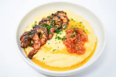 A colorful and appetizing plate of Spicy Grilled Shrimp and Grits featuring succulent, charred shrimp atop creamy, golden grits, complemented by a vibrant cherry tomato confit garnished with fresh herbs.