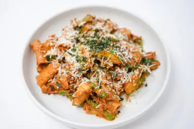 A plate of Rigatoni with Asparagus, Zucchini, and Sun-Dried Tomato Pesto, featuring al dente pasta mixed with bright green asparagus and zucchini, all coated in a vibrant red pesto, sprinkled with grated Parmesan and fresh parsley.