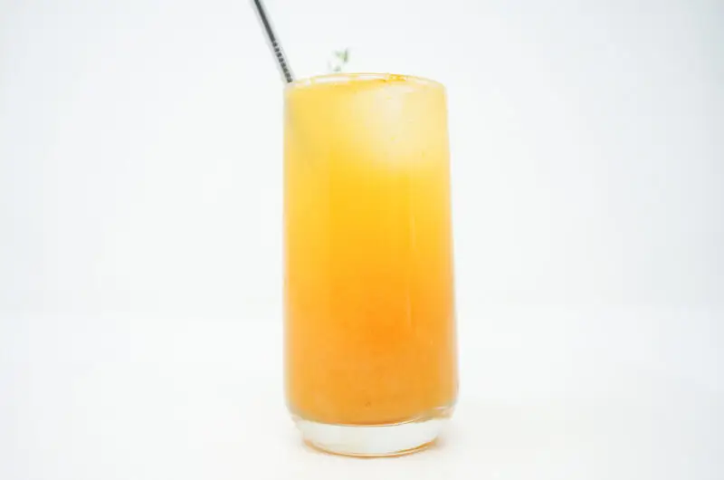 A tall glass of Peach Thyme Bellini, featuring layers of golden peach puree and effervescent Prosecco, garnished with a fresh thyme sprig for a sophisticated touch.