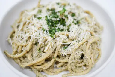 A plate of Wild Mushroom Pesto Bucatini, showcasing thick strands of pasta generously coated in a green, herby wild mushroom pesto, garnished with grated Parmesan and fresh parsley, offering a rustic yet elegant presentation.