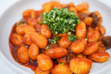 A sumptuous serving of Braised Oxtail Gnocchi with Lemon Gremolata, showcasing tender oxtail and fluffy gnocchi coated in a rich, glossy tomato sauce, garnished with bright green, freshly chopped gremolata.