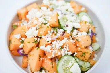 A colorful Mediterranean Cantaloupe Melon Salad, featuring bright orange cantaloupe chunks, crisp cucumber slices, sharp red onion slivers, and crumbled feta cheese, all sprinkled with fresh green mint and drizzled with a lemon-honey dressing, served in a white bowl.