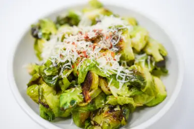A vibrant dish of Lemon Parmesan Roasted Brussels Sprouts, showcasing golden-browned, crispy sprouts sprinkled with melted Parmesan, fresh lemon zest, and finely chopped garlic, served in an elegant white bowl.