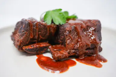 A plate of Southern Style Braised Beef Short Ribs, tender and glistening with a rich, spicy tomato sauce, garnish with fresh parsley, served on a rustic white plate.