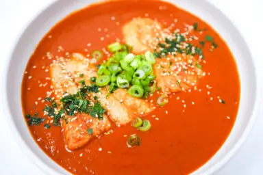 A vibrant plate of Spicy Tomato Sesame Poached Basa, featuring succulent basa fillets in a rich, red tomato sauce sprinkled with toasted sesame seeds, green onion, and garnished with fresh cilantro, served on a sleek white plate.