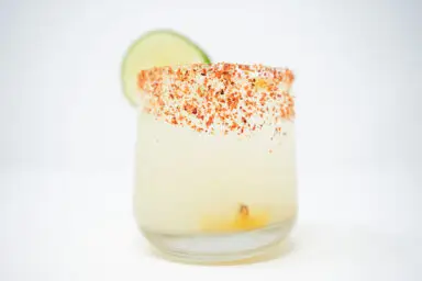 A Sweet Corn Margarita on a clean white background, featuring a glass rimmed with Tajín and topped with roasted corn kernels, highlighted by the vibrant yellow of the corn syrup and a slice of lime for a splash of color.
